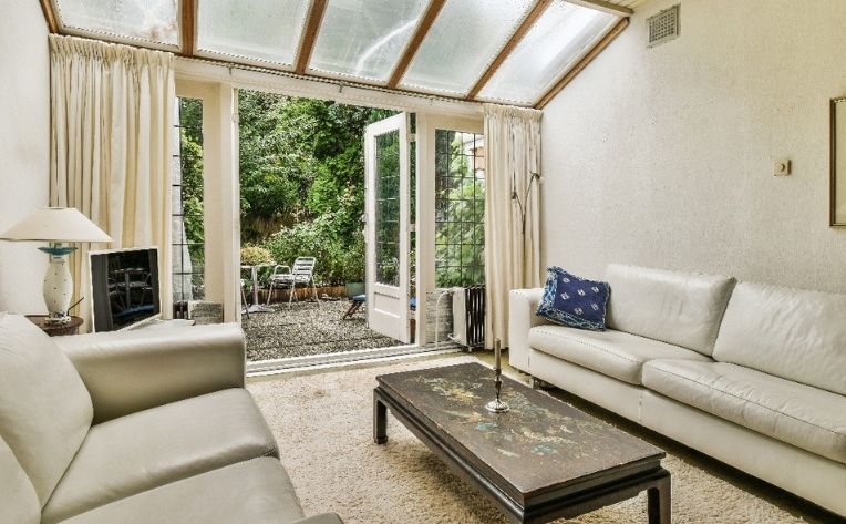 Bright sunroom living area with white leather sofas, glass ceiling, coffee table, and open French doors leading to a garden patio.